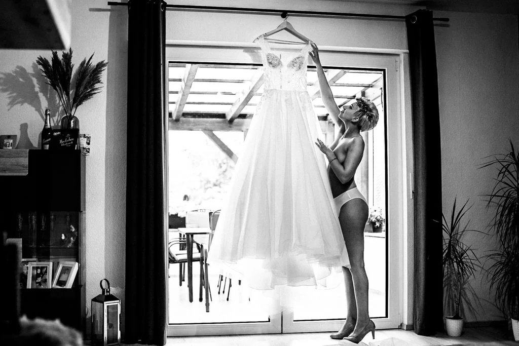 Eine Braut in weißer Wäsche hält ihr Brautkleid ins Fensterlicht
