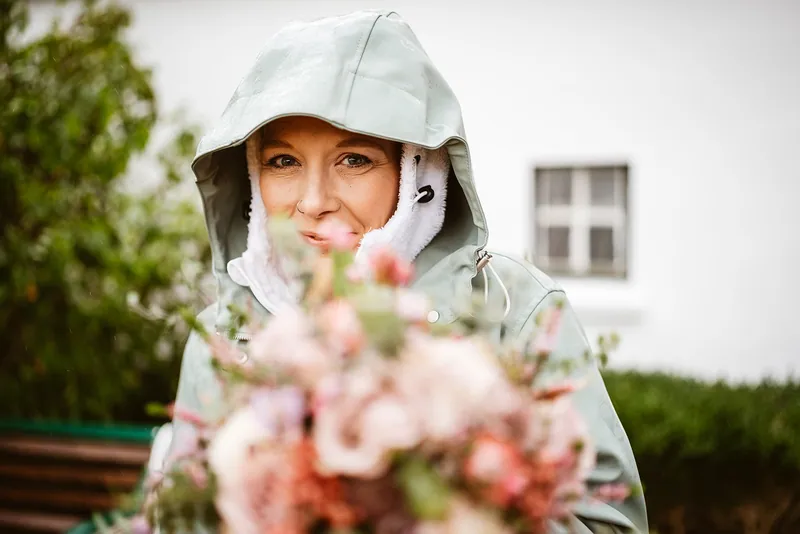 Mel schaut aus ihrer Regenjacke hinter dem Brautstrauß hervor