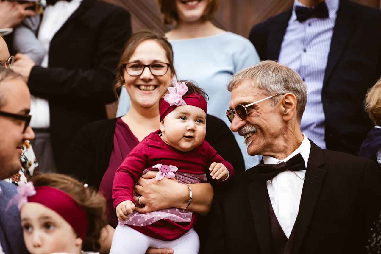 Baby auf dem Arm einer Frau neben einem älteren Mann unter Hochzeitsgästen